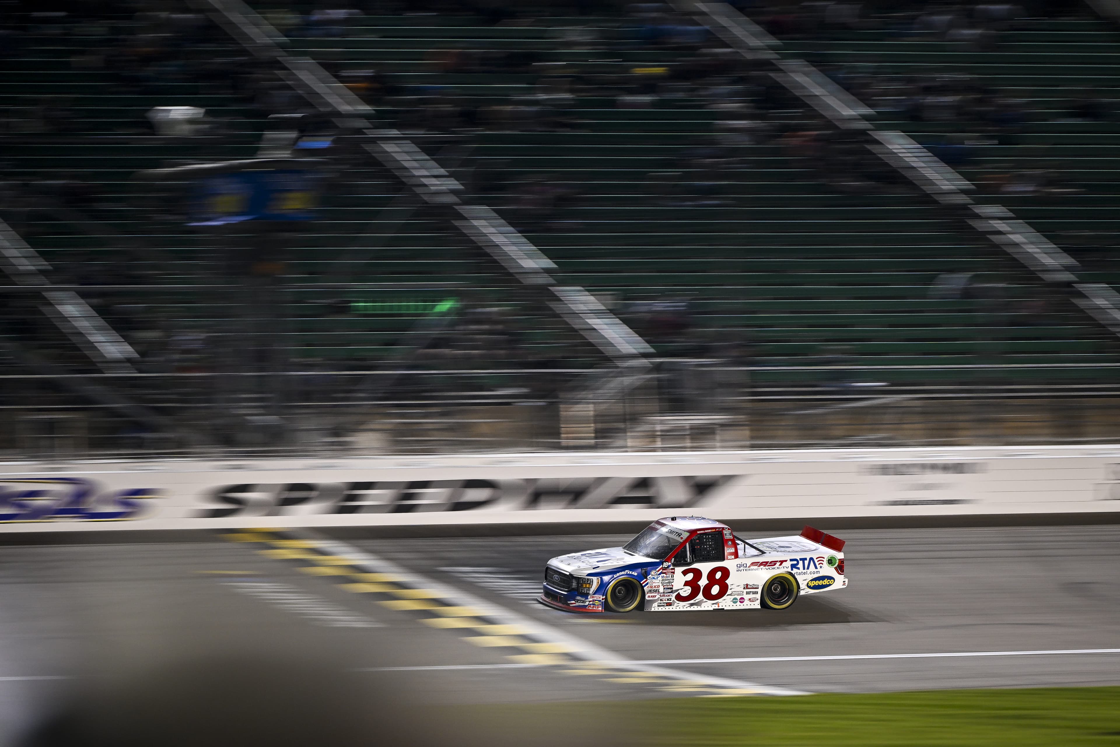 #38 truck at night, passing SPEEDWAY signage, cinematic low-angle shot